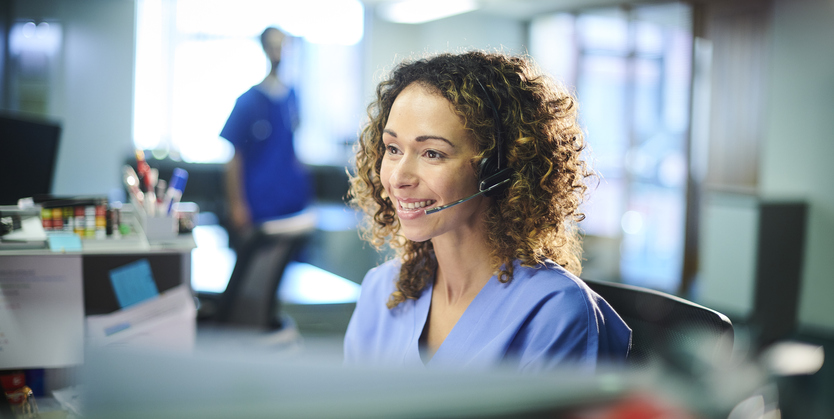 a woman with curly brown hair and scrubs answers the phone at a desk.
